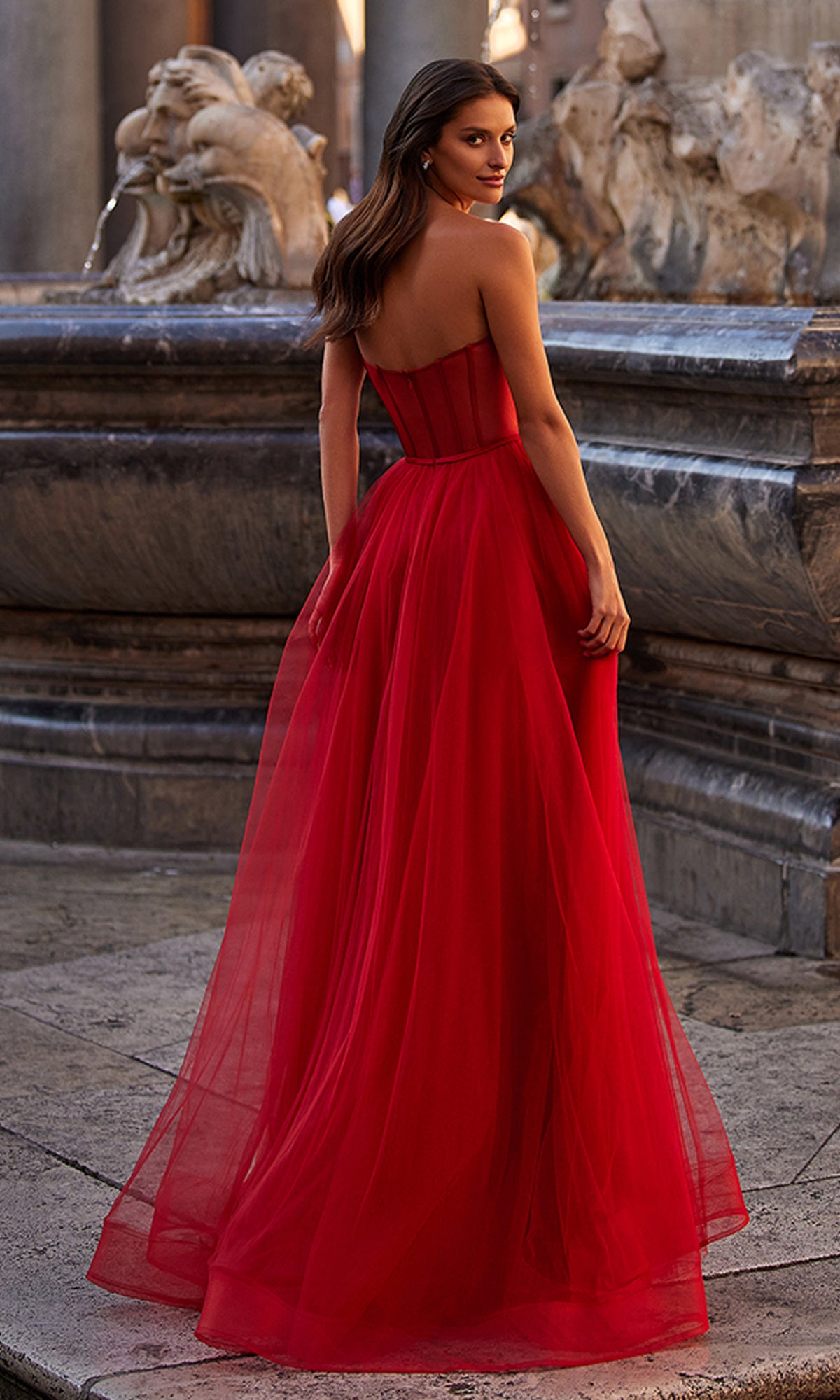 A woman wearing the La Femme 32424 Strapless Long Tulle Prom Dress with a corset-style bodice stands by a stone fountain, glancing over her shoulder.